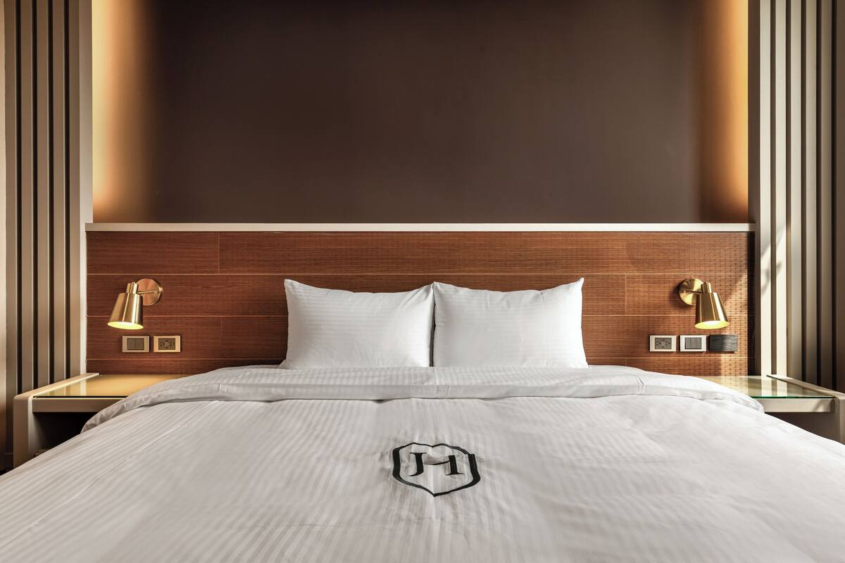 A neatly made bed with a white bedspread and a monogram, two bedside lamps, and a wooden headboard.