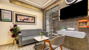 A modern waiting area with a grey sofa, a wooden chair, a small table with a vase, and a framed picture on the wall. - Treebo Arizona (Gandhinagar)