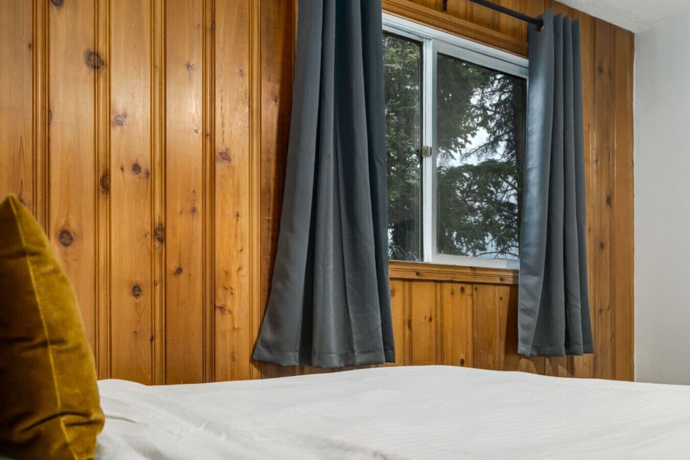 Een kamer met houten lambrisering, een bed met wit beddengoed en grijze gordijnen.
