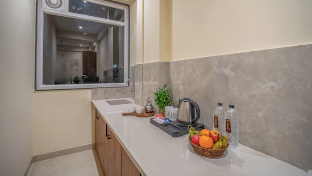 Une cuisine moderne avec une grande fenêtre, un évier, une bouilloire, un panier de fruits et deux bouteilles d’eau posées sur le comptoir.