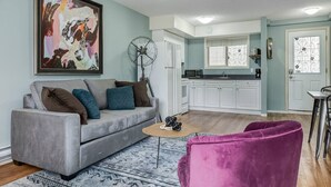 A living room with a grey sofa, a coffee table, and a purple chair. There is a kitchen in the background with white cabinets and a window with decorative ironwork. - Stunning Mountain View Cozy Studio for Small Grp (Canmore)
