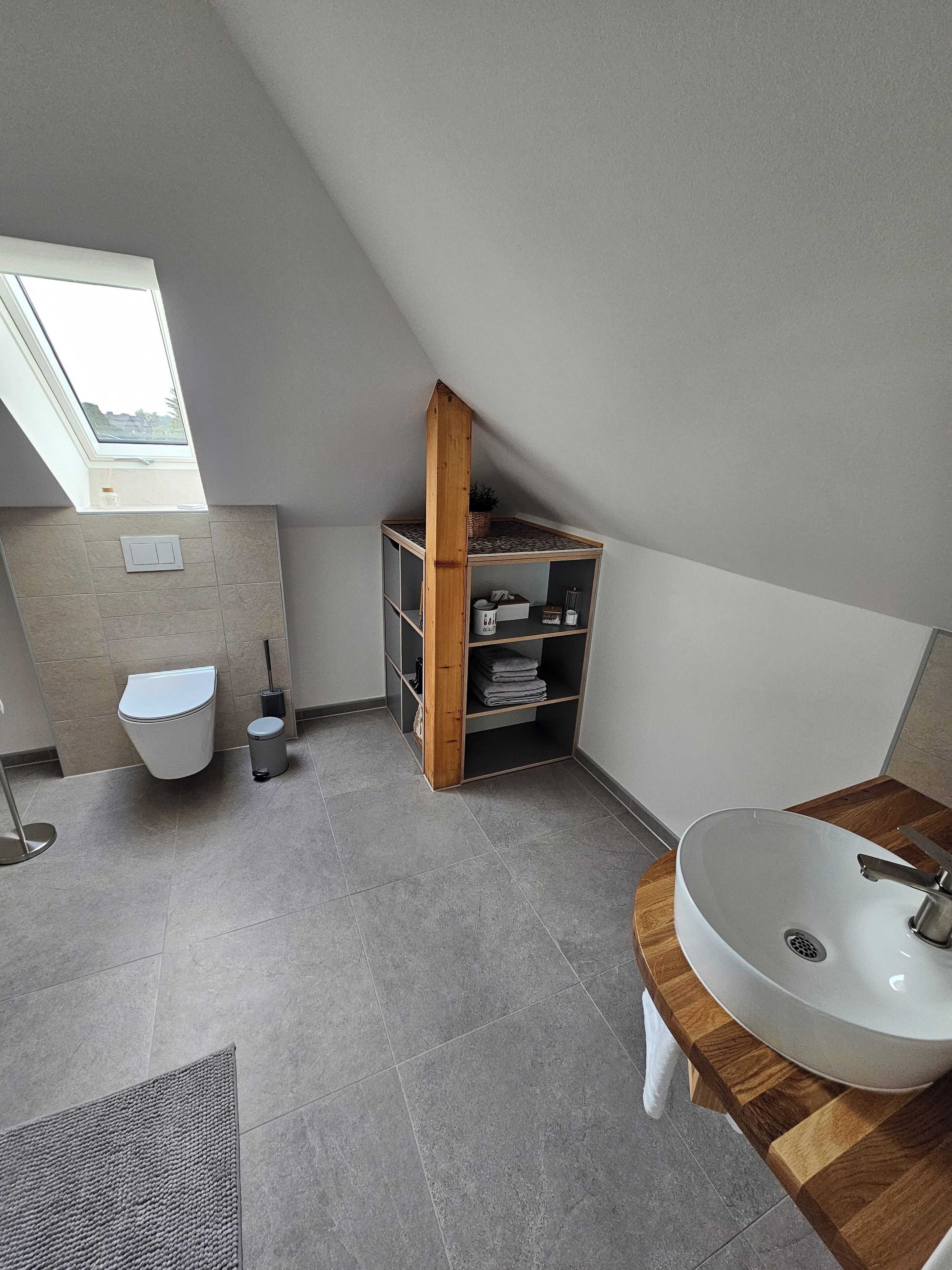 Un baño moderno con tragaluz, una estantería de madera, un inodoro, un cubo de basura y un lavabo con encimera de madera.