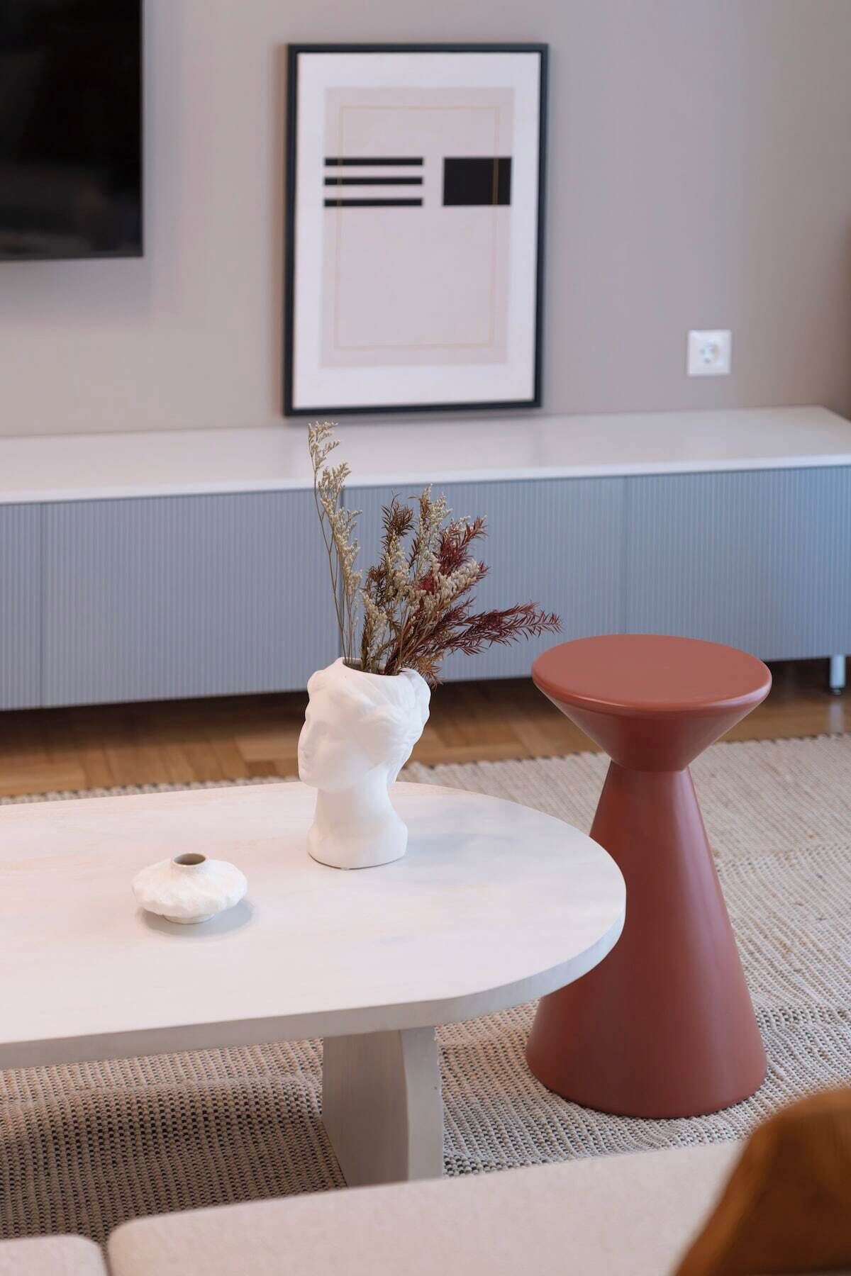 A modern living room with a round white table, a white vase with dried flowers, a small white object, and a red stool.