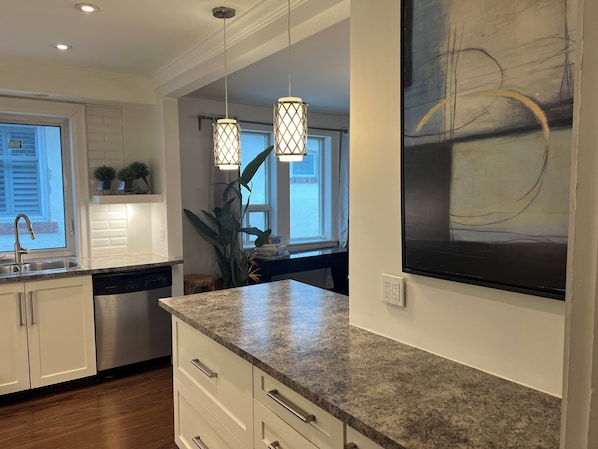 A modern kitchen with white cabinetry, granite countertops, pendant lighting, and a large abstract painting on the wall.
