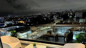 Un balcón con mobiliario de mimbre, una pequeña mesa con una planta y vistas a un paisaje urbano nocturno.