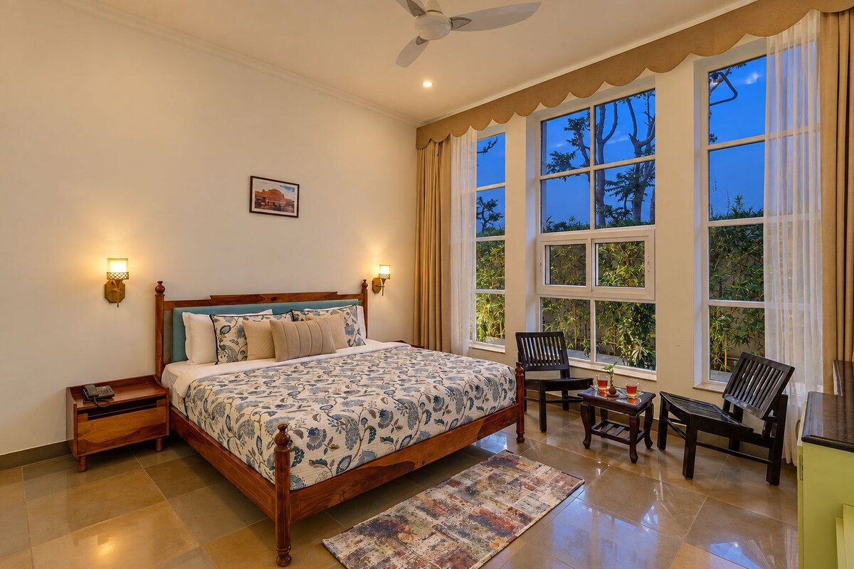 A bedroom with a large bed, two bedside tables, a chair, a small table with a drink, and a view of trees through the window.