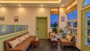 A cozy lounge area with a sofa, armchairs, and a coffee table. There is a door with a number sign, a window with a view, and a potted plant.