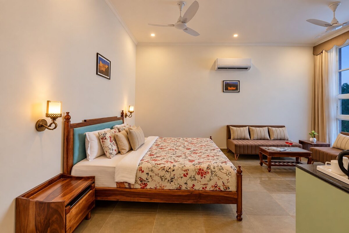 A hotel room with a bed, a sofa, a coffee table, and a desk with a chair. There are two ceiling fans, an air conditioning unit, and a window with curtains.