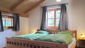 Ein hölzernes Bett mit grüner Bettwäsche in einem Zimmer mit Holzbalken an der Decke, einem Fenster mit Vorhängen und einer Nachttischlampe.