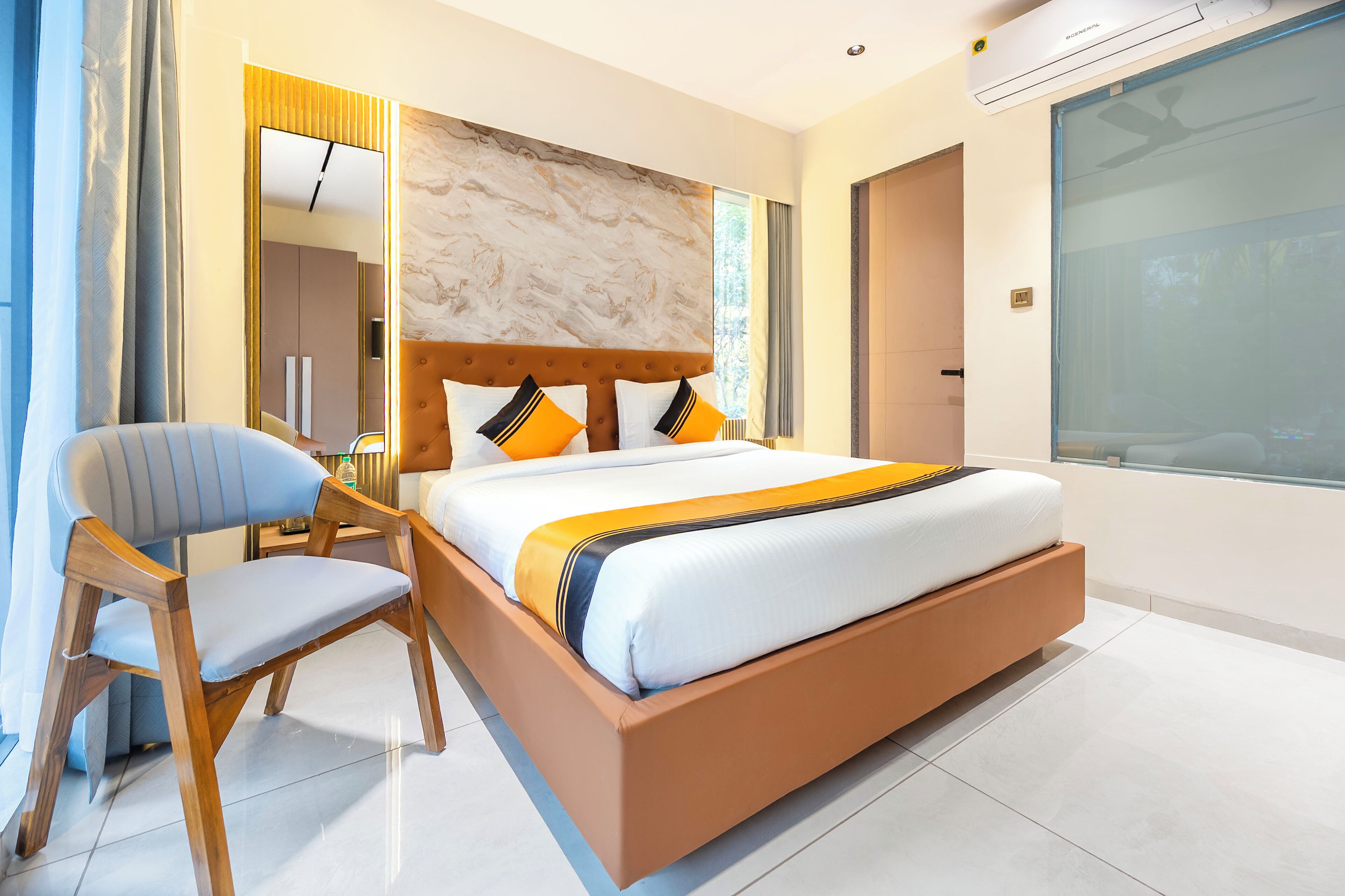 Una habitación de hotel moderna con una cama grande, una silla de madera, un espejo y un ventanal amplio.