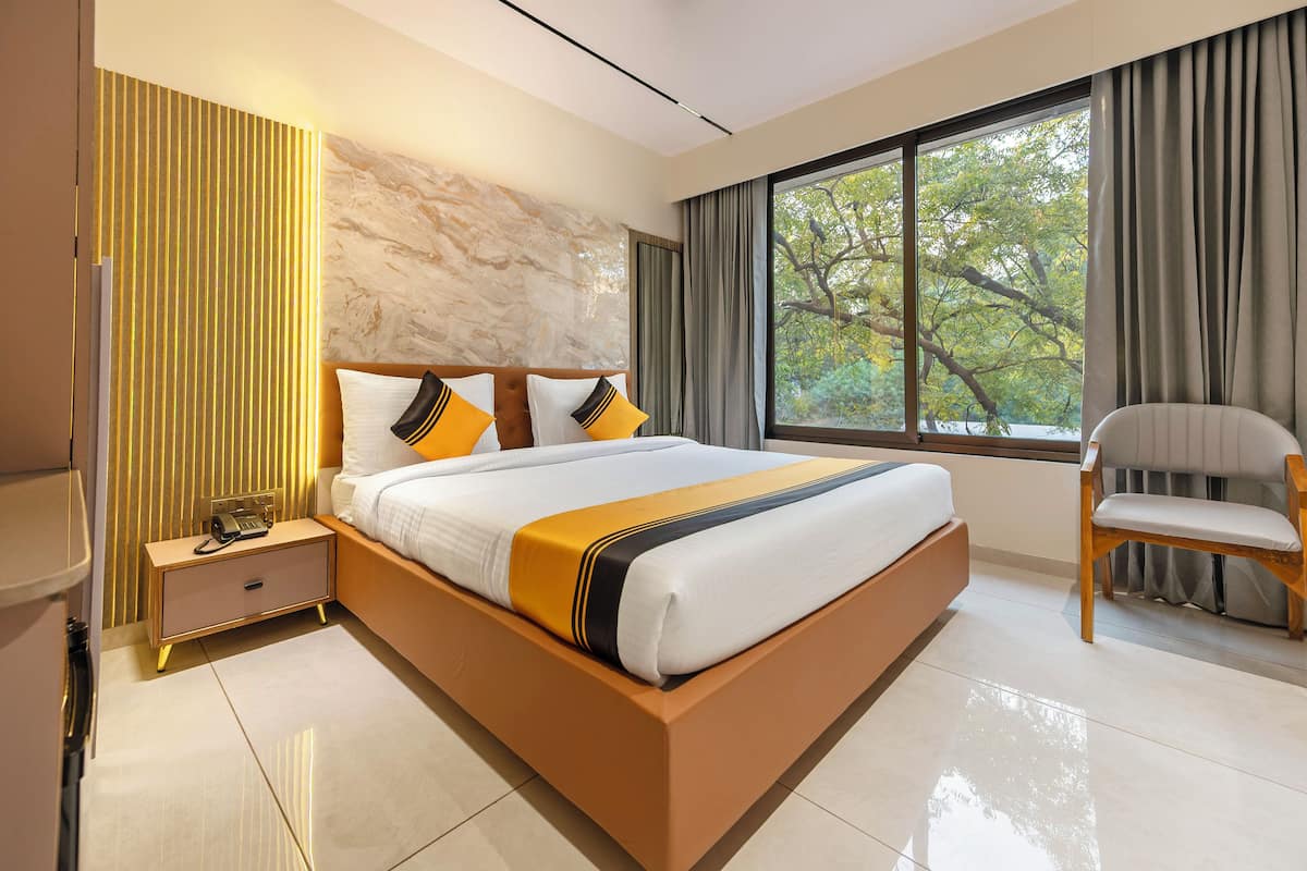 Una habitación de hotel moderna con una cama grande, una mesita de noche, una silla y vistas a árboles por la ventana.
