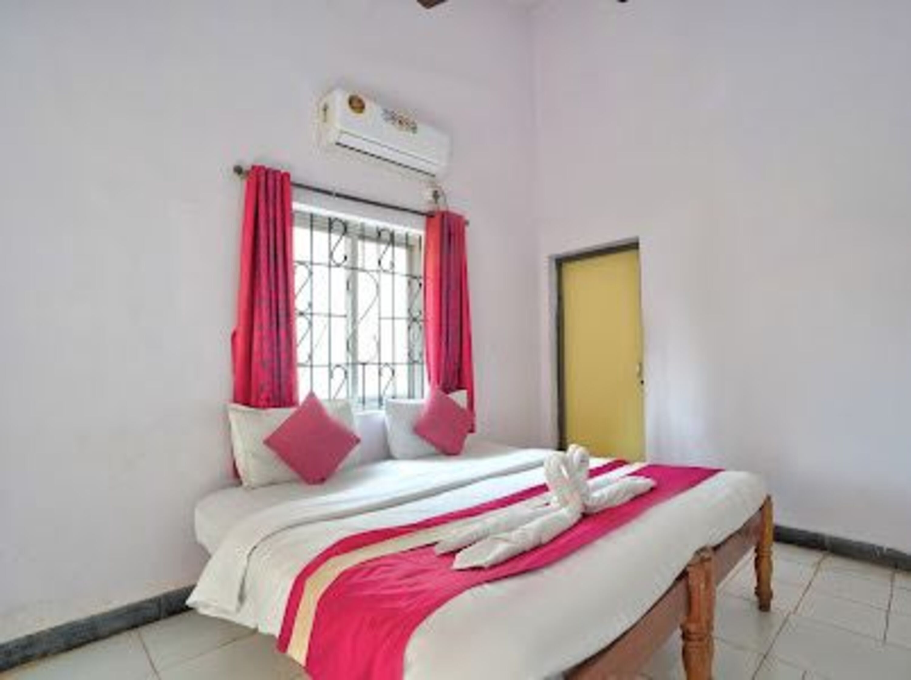 A bedroom with a bed, a window with metal bars, a yellow door, and an air conditioning unit on the wall.