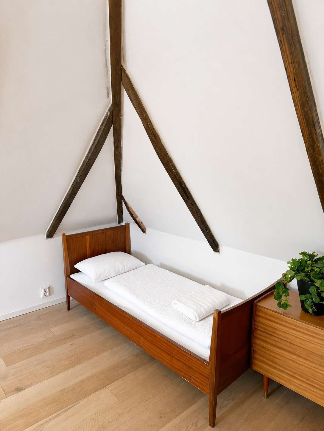 A single bed with a wooden frame and white bedding in a room with sloped ceilings and exposed wooden beams.