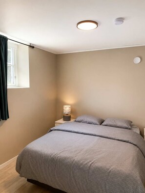 Een slaapkamer met een bed, een nachtkastje met lamp, een raam met gordijnen en een plafondlamp.