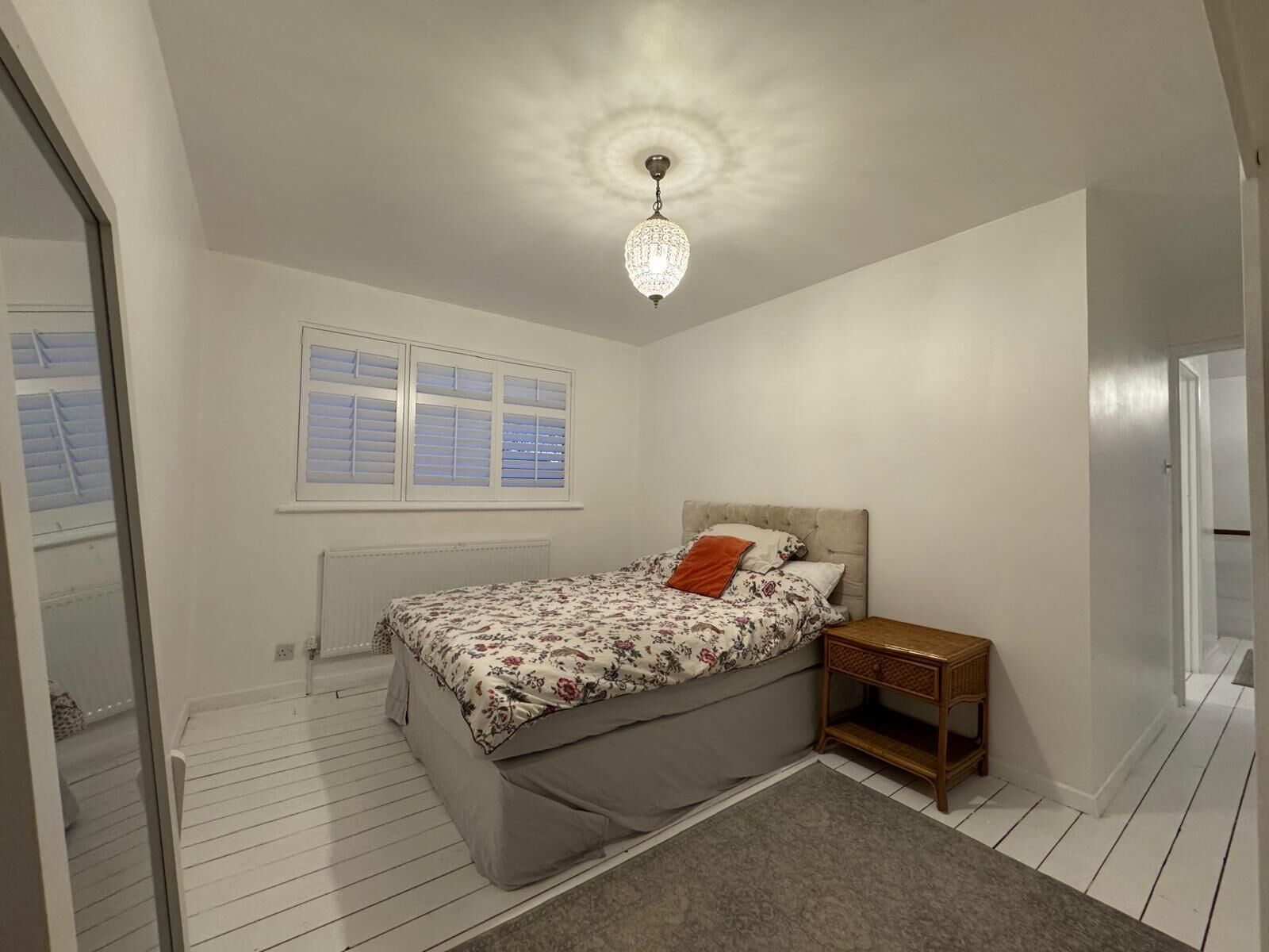 A bedroom with a bed, a nightstand, a window with blinds, a radiator, and a chandelier.