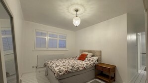 Een slaapkamer met een bed, een nachtkastje, een raam met jaloezieën, een radiator en een kroonluchter.