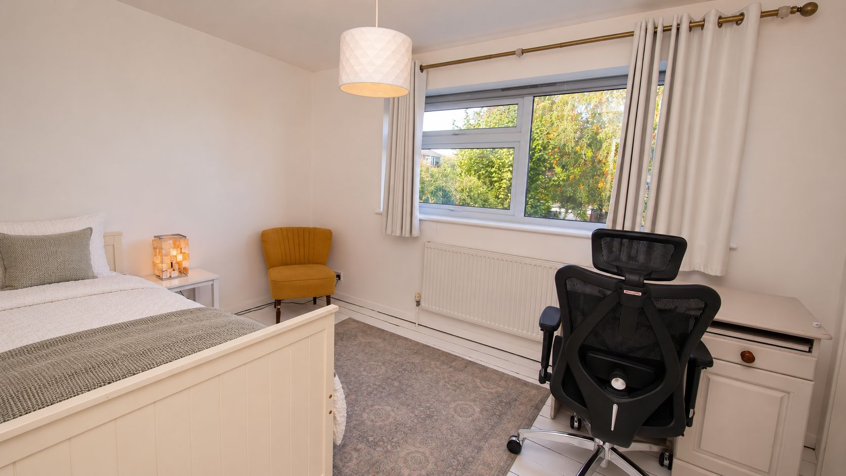 A bedroom with a bed, a chair, a desk with a chair, a lamp, and a window with curtains.