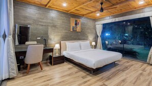 A modern bedroom with a wooden ceiling, a large bed, a desk with a chair, and a view of the outdoors through a glass door. - Eco Holiday Resorts (Indore)