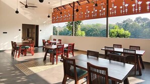 A spacious dining area with multiple wooden tables and chairs, ceiling fans, and large windows offering a view of greenery. - Vaigayi Ayurvedic Resort (Kunnathunad)