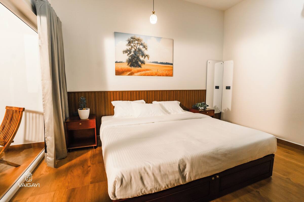A neatly arranged bedroom with a large bed, a bedside table with a lamp, a potted plant, and a window with curtains.
