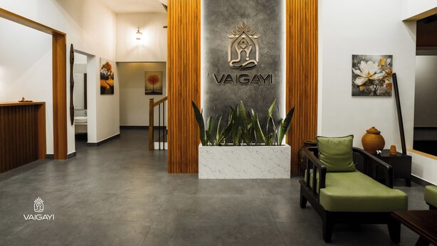 A reception area with a modern design, featuring a reception desk, a seating area with a sofa and armchairs, a potted plant, and a wall-mounted logo. - Vaigayi Ayurvedic Resort (Kunnathunad)