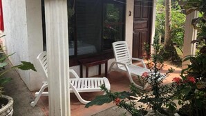 Un espace extérieur abrité, aménagé avec deux chaises en plastique blanc et une petite table, entouré de plantes en pot et d’une porte en bois.