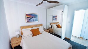 Una habitación de hotel con una cama, mesitas de noche, un ventilador de techo, un espejo grande y un cuadro en la pared.