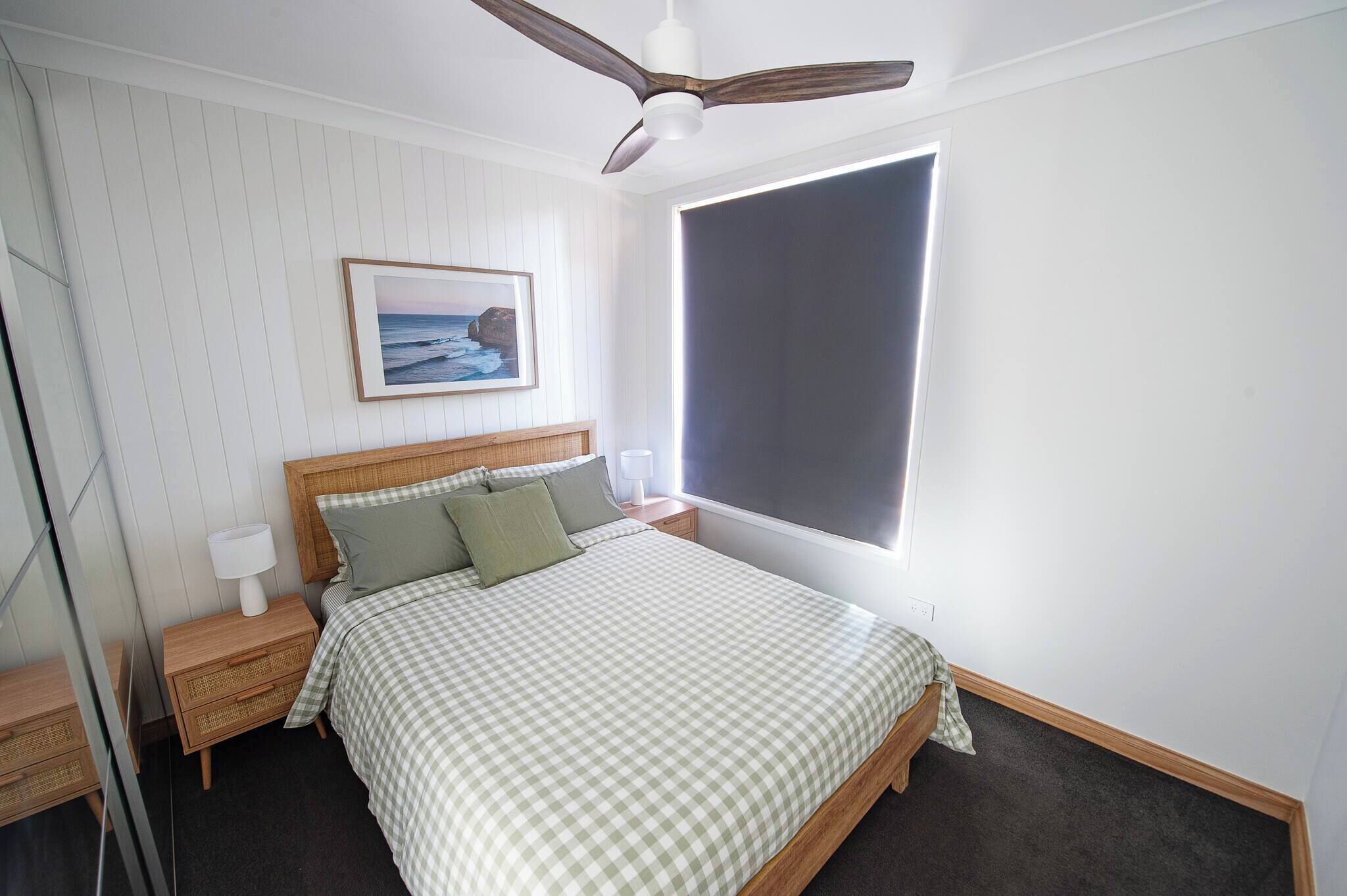 Une chambre bien rangée, avec un lit, des tables de chevet, un ventilateur de plafond, une fenêtre et un tableau encadré au mur.