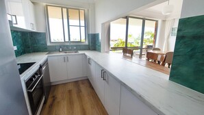 Una cocina moderna con armarios blancos, salpicadero de mármol y piso de madera. Hay un ventanal que proporciona luz natural y vistas al exterior.