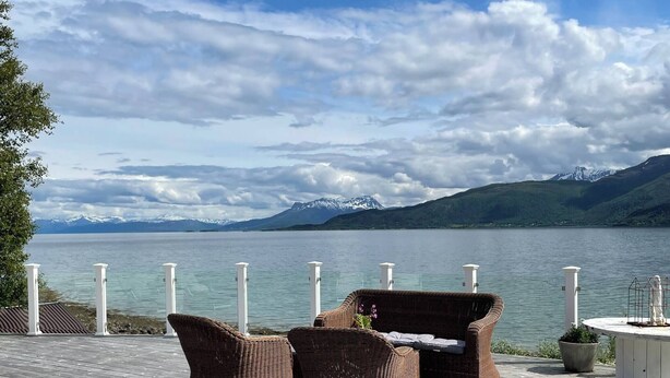 Een houten terras met rieten meubels en een kleine tafel, met uitzicht op een meer met bergen op de achtergrond.