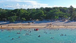 Ein Strand mit klarem türkisblauem Wasser, Menschen beim Schwimmen und Sonnenbaden, und eine Baumreihe im Hintergrund.