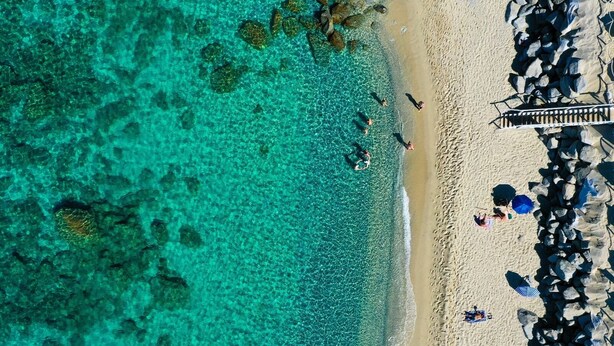 Vue aérienne d’une plage aux eaux turquoise et limpides, bordée d’un rivage sablonneux et d’une jetée agrémentée de parasols.