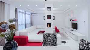 A modern living room with a white sofa, red accent pillows, a black and white patterned ottoman, a chandelier, and a flat-screen TV on a white stand. - L'onde du Lac - chic apartment with private suite prés aéroprt et cliniques (Tunis)