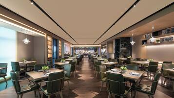 A modern dining area with multiple tables and chairs, a central bar, and a view of the kitchen in the background.