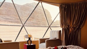 A bedroom with a large bed, a view of a desert landscape through a geometric window, and a cozy seating area with a small table and a potted plant. - Wadi Rum Main Road Visitor Center (Wadi Rum Village)