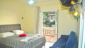Un dormitorio con cama, sofá, mesa y balcón con vistas a edificios y zonas verdes.