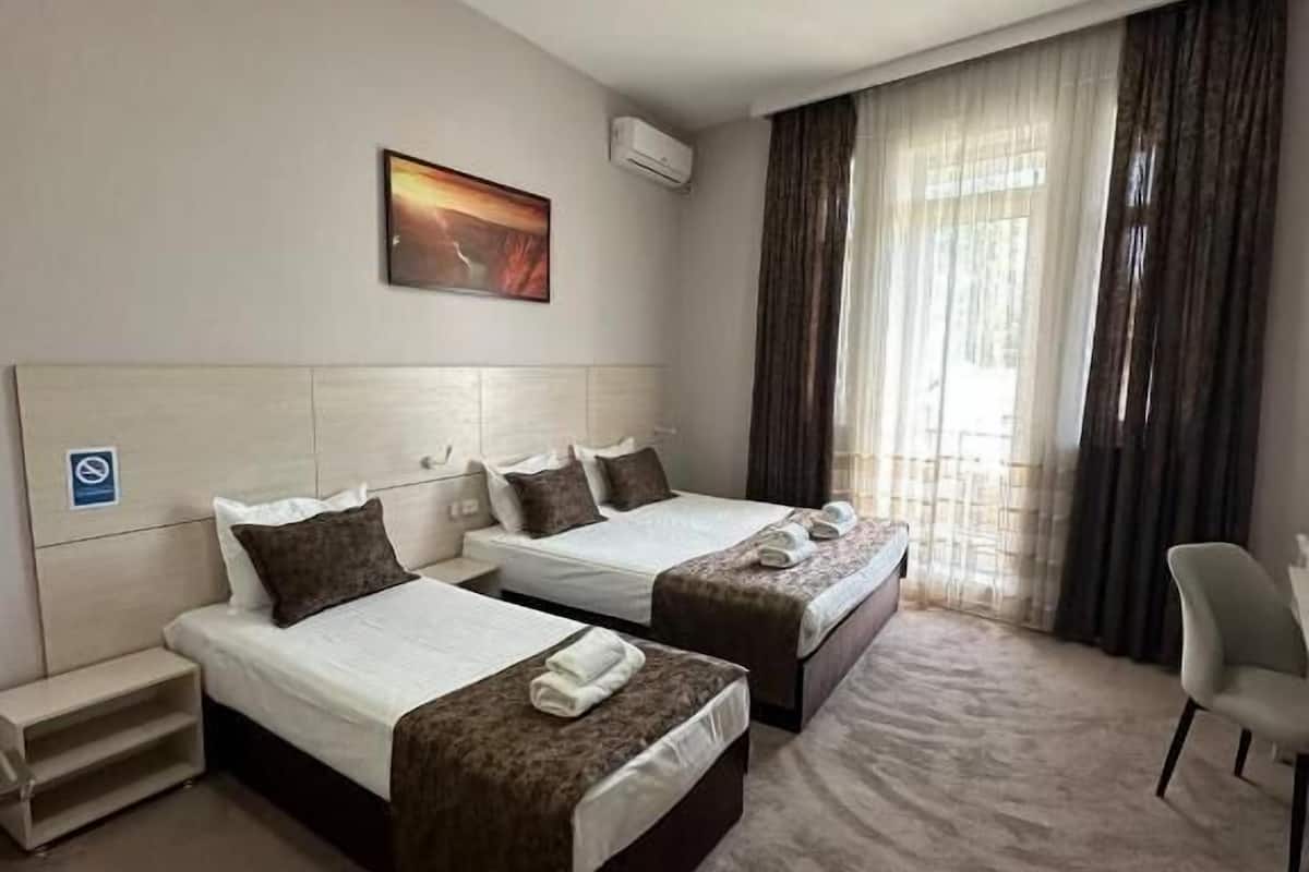A hotel room with two beds, a desk, a chair, a painting, and a window with curtains.