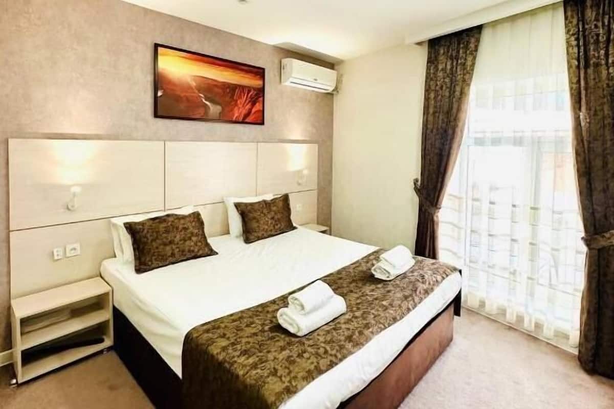 A hotel room with a large bed, a nightstand, a wall-mounted air conditioner, a painting, and a window with curtains.
