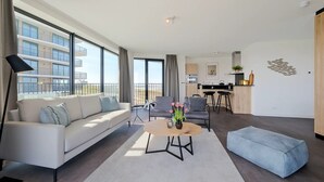 Ett modernt vardagsrum med en soffa, ett soffbord och en pall. Här finns också en köksdel med ett matbord och stolar. Stora fönster ger utsikt över byggnadens fasad.