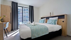 Ett modernt hotellrum med en stor säng, ett skrivbord och utsikt över en byggnad genom tunna, genomskinliga gardiner.