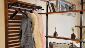 Quarto com escada de madeira, um cabideiro com jaquetas, uma estante com livros e um quadro, e uma pequena mesa com um vaso.