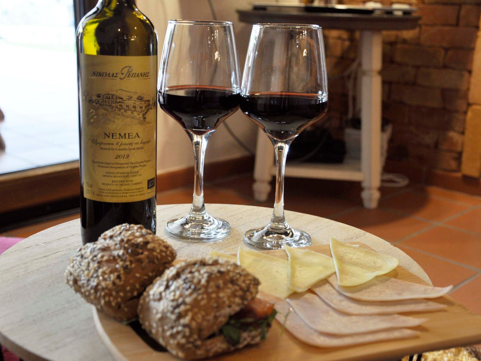 A bottle of NEMEA wine, two wine glasses, and a wooden board with sandwiches and cheese.