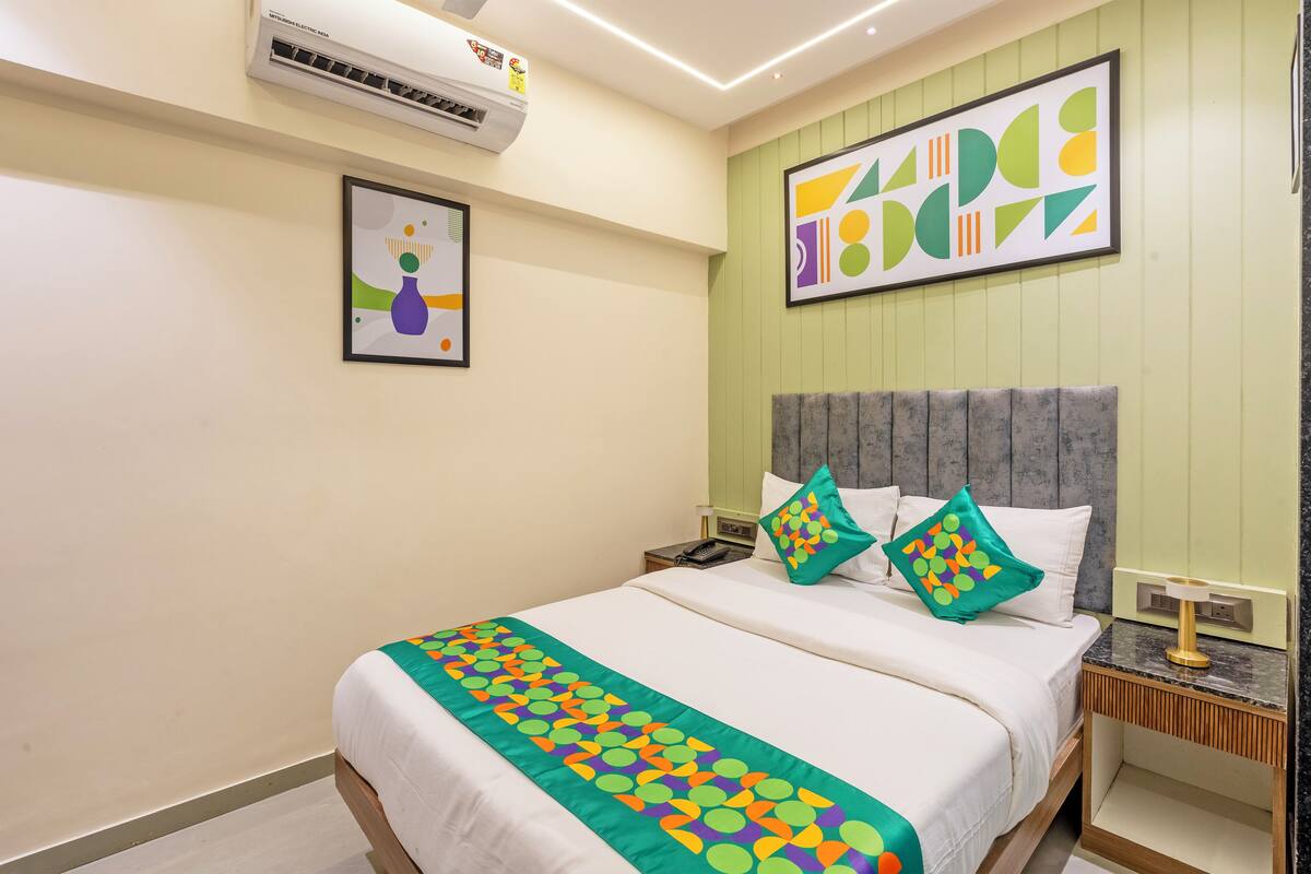 A hotel room with a bed, a nightstand, a wall-mounted air conditioner, and a framed picture.