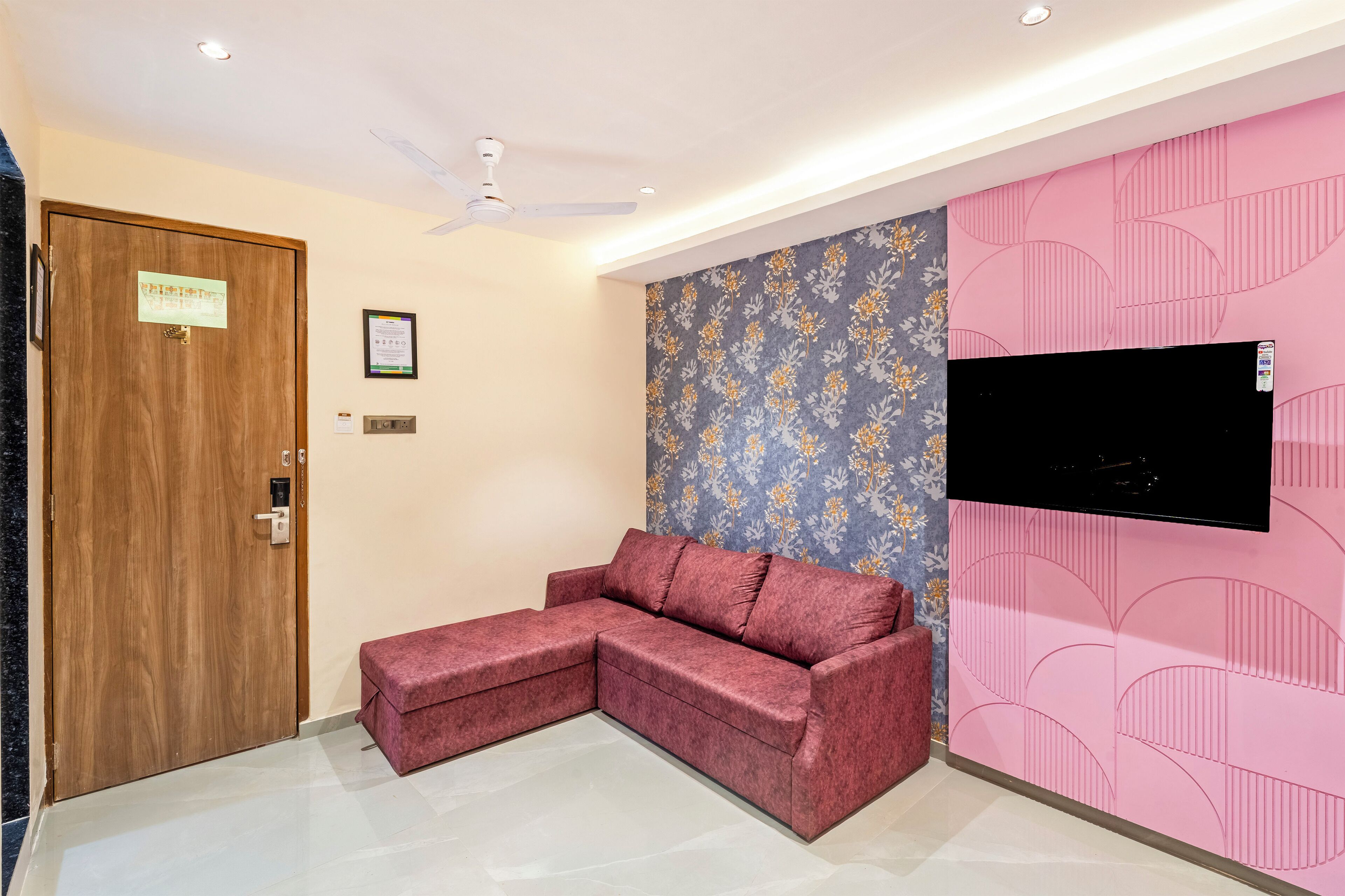 A room with a wooden door, a ceiling fan, a pink wall with a TV mounted on it, and a red sofa.