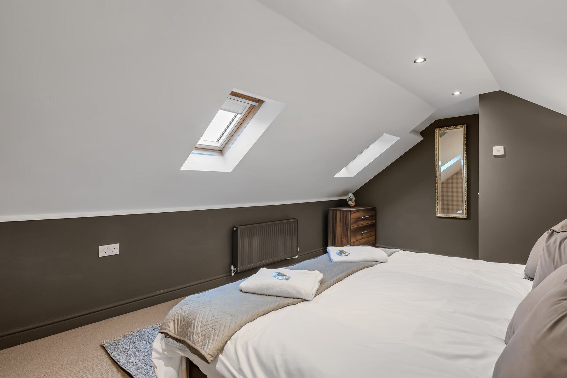 Een slaapkamer met dakraam, radiator, nachtkastje en spiegel.