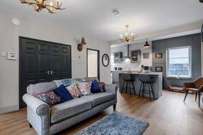 A modern living room with a grey sofa, a small rug, a kitchen with a bar area, and a large window. - Pools Cottage 1 Game Keepers Eco-Friendly Retreat (Kenilworth)