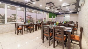 A dining area with multiple tables and chairs, large windows, and ceiling fans. - Treebo Arya Palace With Swimming Pool (Shakti Enclave)