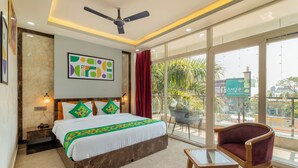 A hotel room with a large bed, a ceiling fan, a balcony with a chair, and a view of the city. - Treebo Arya Palace With Swimming Pool (Shakti Enclave)