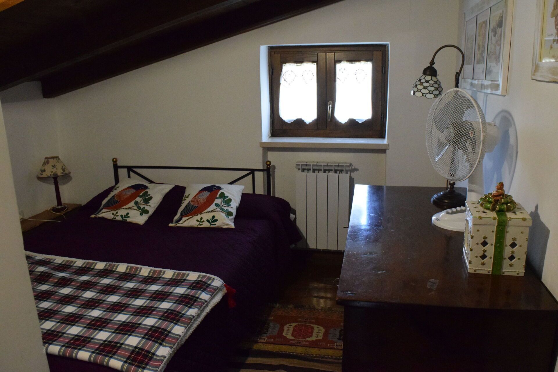 Un dormitorio con cama, mesita de noche, cómoda, ventana, radiador, ventilador y una pequeña mesa con un florero.