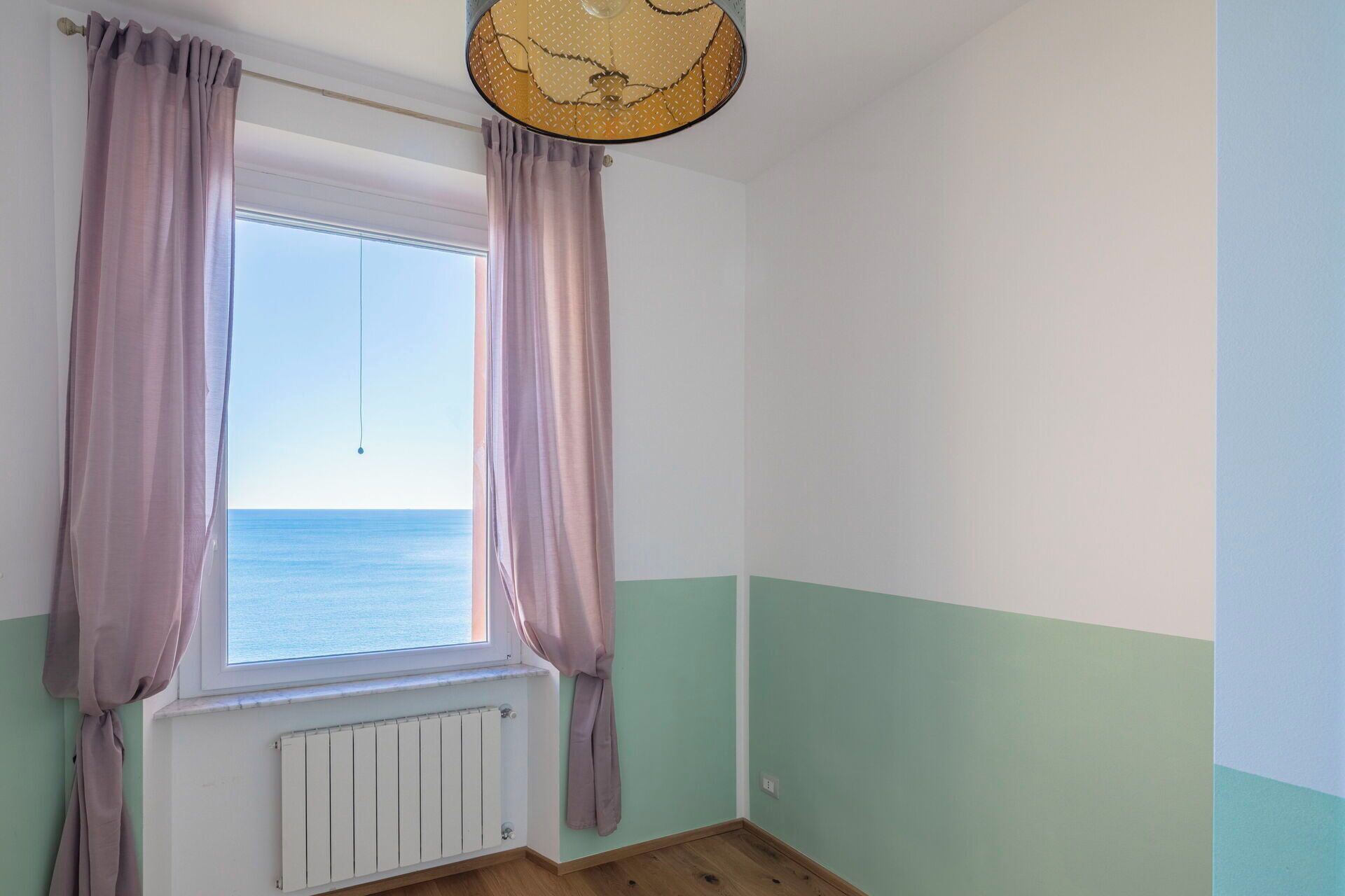 A room with a window overlooking the sea, a radiator below the window, and light green walls.